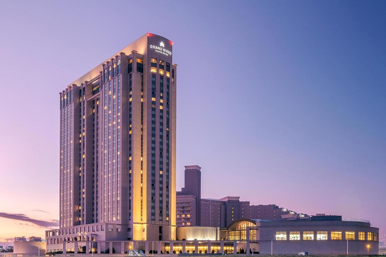 Grand Nikko Tokyo Daiba Hotel Exterior photo The image shows a tall hotel building, likely a high-rise, which is illuminated against a twilight sky. The facade features a mix of modern architectural elements, with large windows and a prominent sign at the top. Adjacent to the hotel, there are a