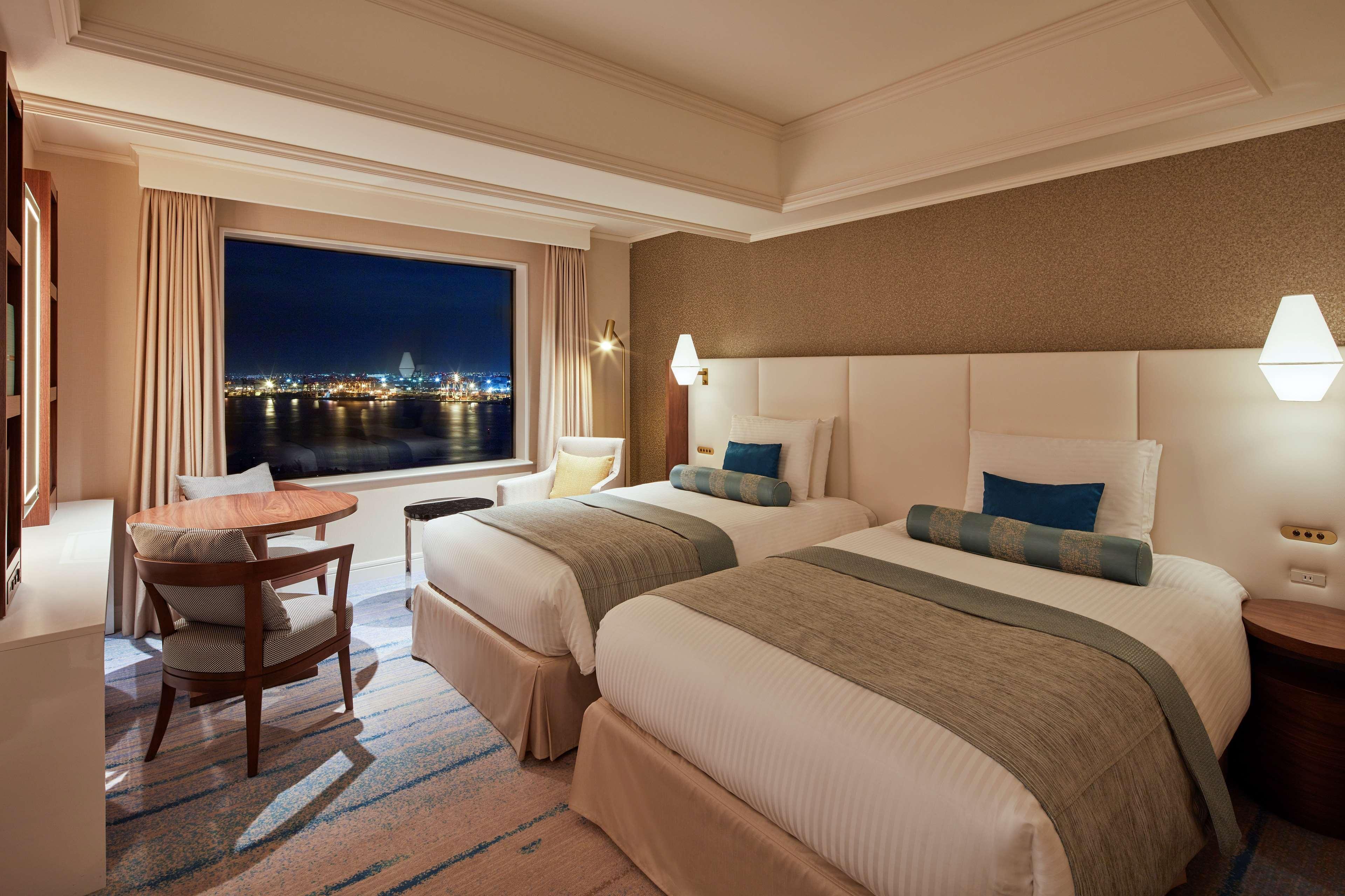 Grand Nikko Tokyo Daiba Hotel Exterior photo The photo shows a cozy hotel room featuring two twin beds with light-colored bedding. The room is elegantly decorated with neutral tones and has a soft blue accent on the pillows. There's a small wooden table and chairs positioned near a large window