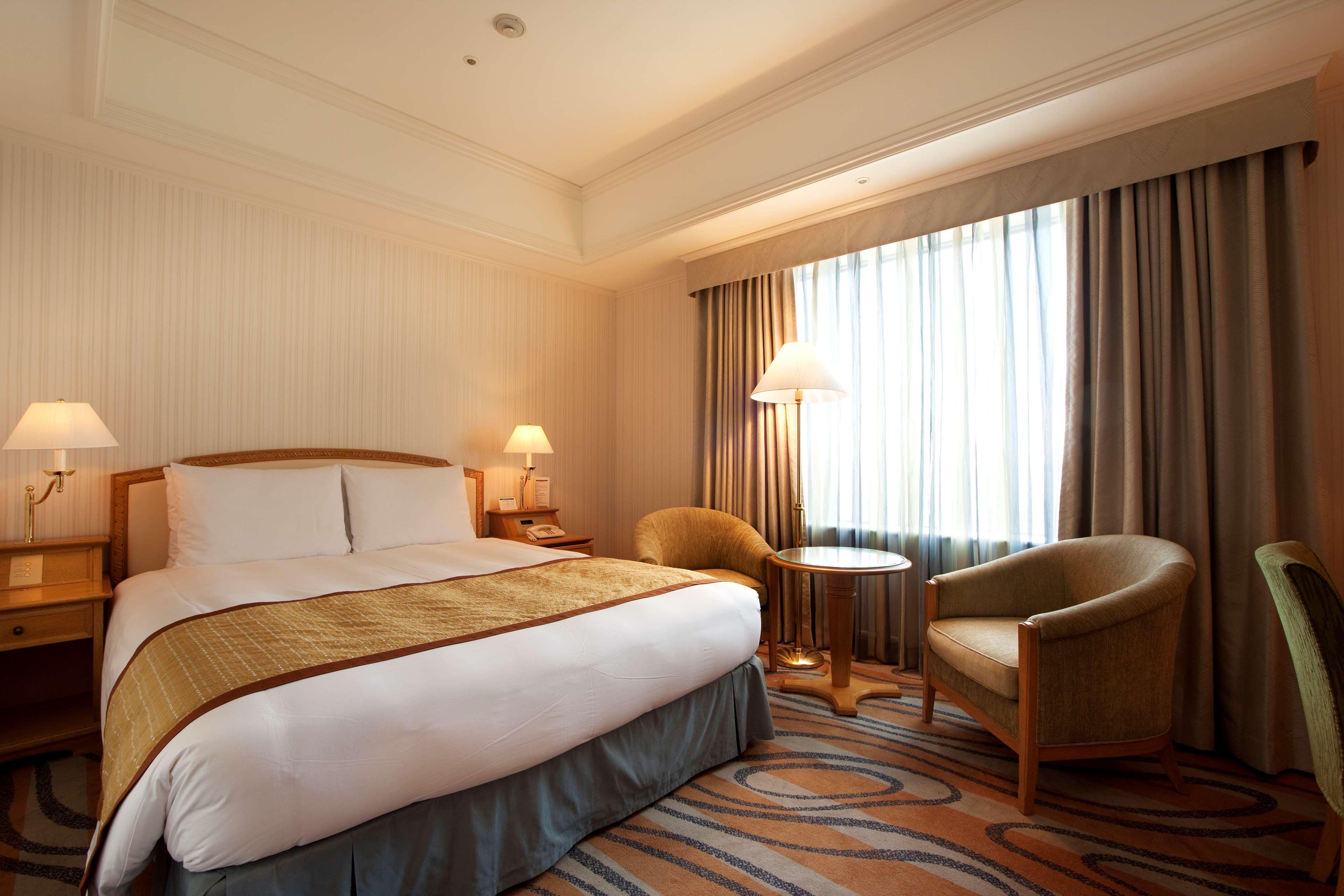 Grand Nikko Tokyo Daiba Hotel Exterior photo The photo shows a hotel room with a comfortable setup. There is a large bed with a white duvet and a decorative brown throw blanket at the foot. On either side of the bed, there are bedside tables with lamps. A set of two cozy armchairs and a small r