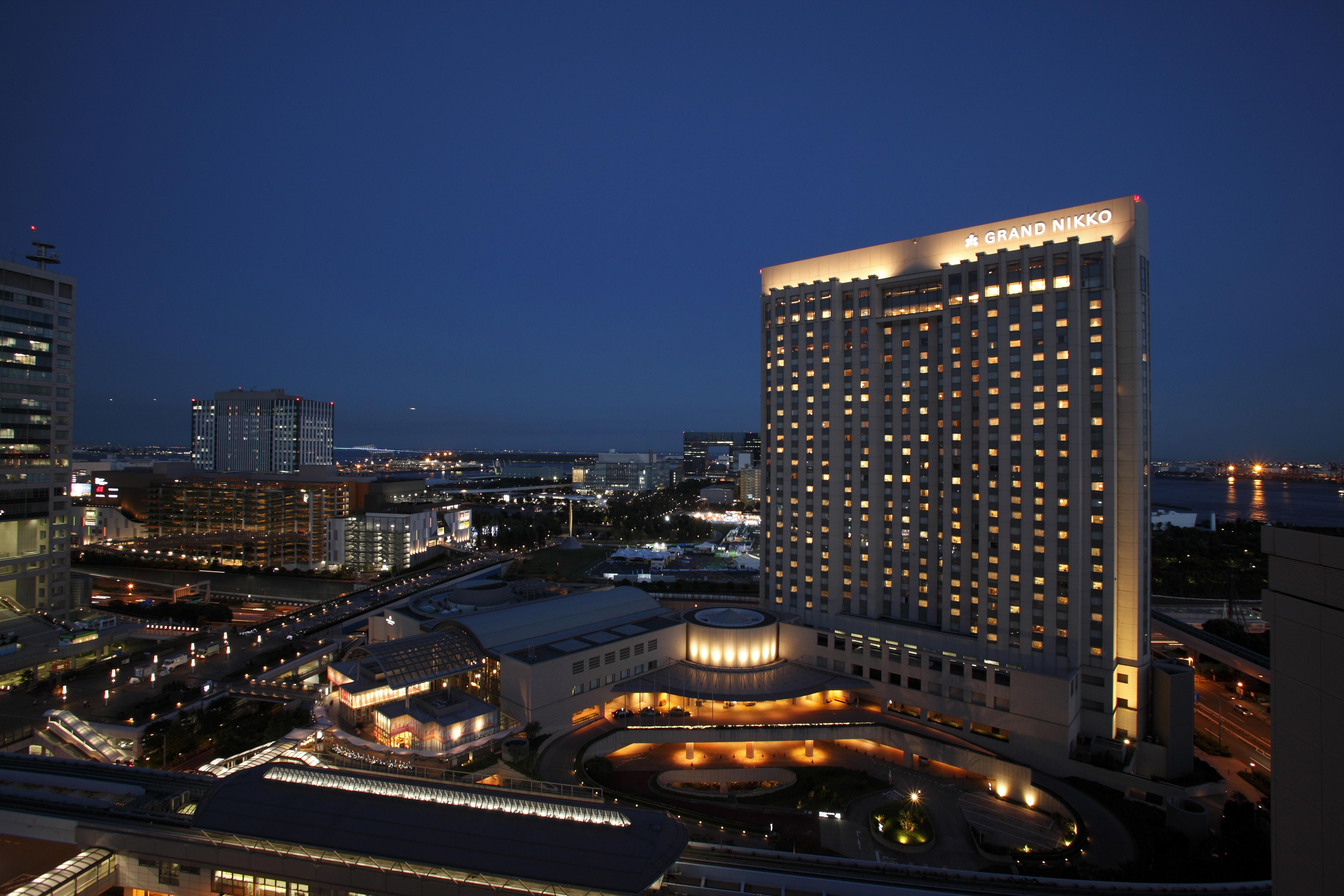 Grand Nikko Tokyo Daiba Hotel Exterior photo The image shows a modern hotel at night, illuminated with numerous lights. The building has a sleek design with a prominent facade featuring many windows that reflect the city lights. Surrounding the hotel are other structures, likely part of a large