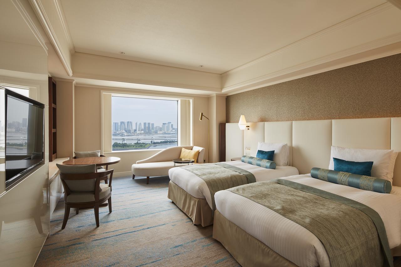 Grand Nikko Tokyo Daiba Hotel Exterior photo The photo shows a well-furnished hotel room featuring two beds covered with light-colored blankets. There is a comfortable chaise lounge near the window, accompanied by a small wooden table and a chair. A flat-screen television is mounted on the wall